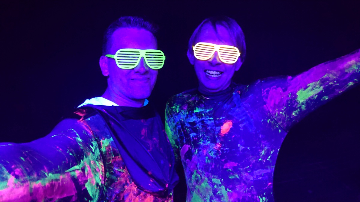 Das Foto zeigt zwei DJs mit mit UV-Farbe beschmierten Oberteilen unter UV-Licht.