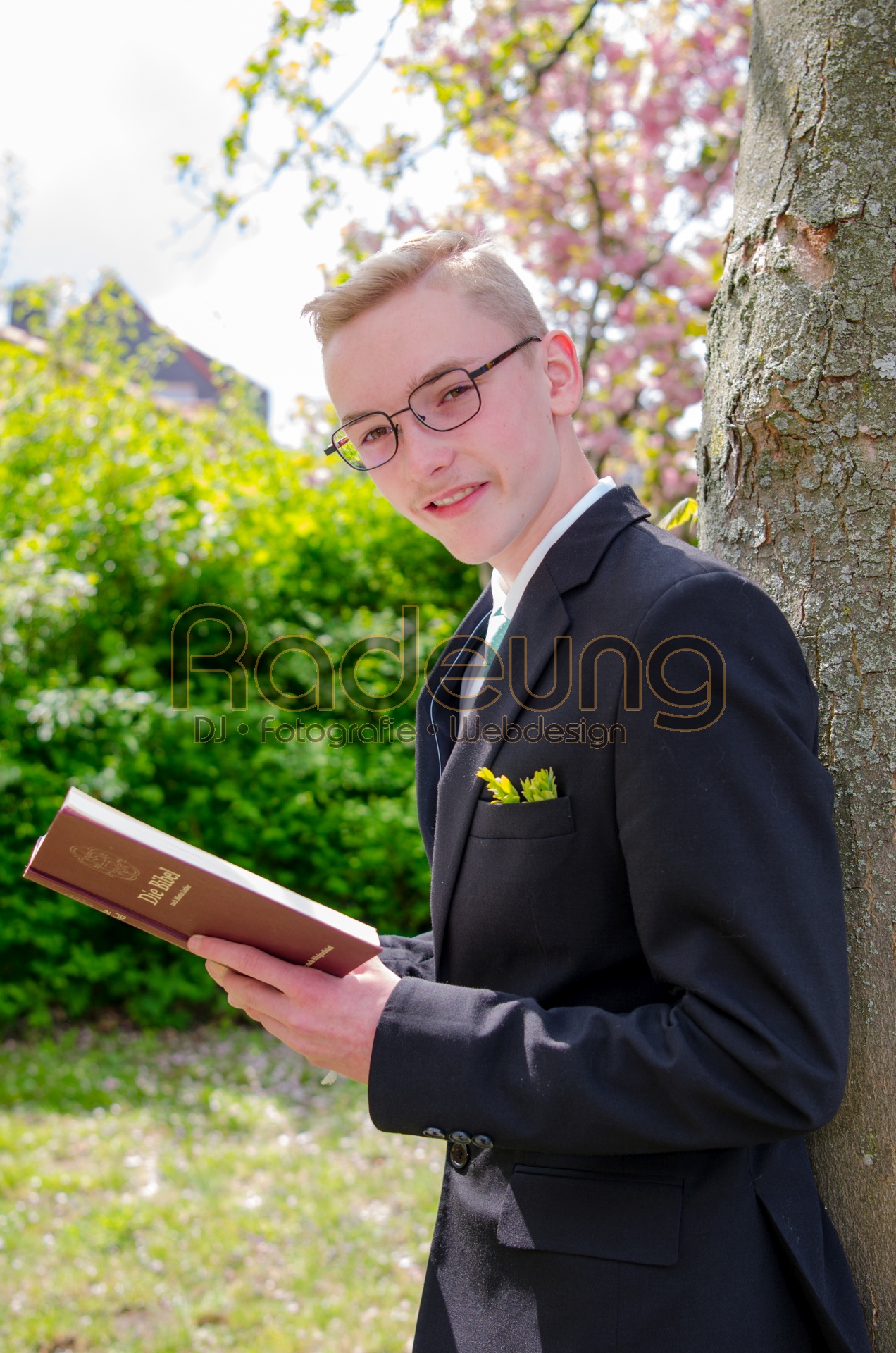 Foto eines Konfirmanden, der an einem Baum lehnt und eine Bibel in der Hand hat